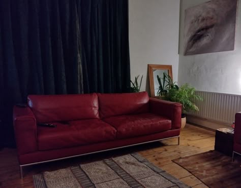 A long, low-backed, red leather sofa stands on a wooden floor. Dark green curtains behind. Abstract art to the right on the wall. A mirror and houseplants in the corner. A Turkish-style rug in the foreground. Red Sofa Leather, Maroon Leather Couch, Red Leather Sofa Living Room Ideas, Vintage Red Couch, Burgundy Leather Couch, Red Leather Sofa Living Room, Red Leather Couch Living Room, Red Leather Couch, Maroon Sofas