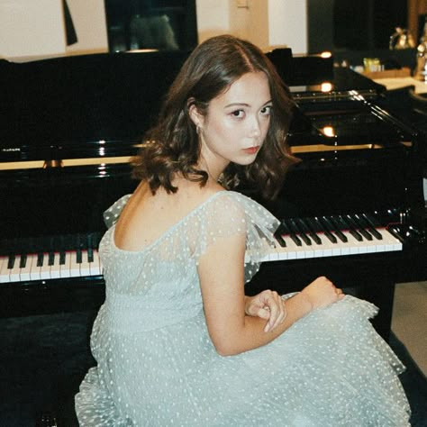 Laufey, a petite woman with short brown hair sits with her back turned to the camera. She is seated at a piano bench in front of a shiny grand piano, and looking over her shoulder in a nonchalant, casual manner. She is appears to be backstage before a concert, and is performance ready in a ruffly light blue gown. Laufey Concert, I Love Laufey, Laufey Lin, Concert Attire, Falling Behind, Concert Pictures, Fav Artist, Fav Music, Do Not Be Afraid