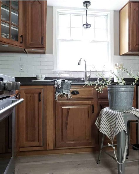 White subway tile backsplash kitchen is equipped with wood cabinets and black countertops. Black and glass pendant lighting hangs in front of kitchen window over single handle faucet. A rustic metal bucket rests upon a matching stool. Oak Cabinets White Quartz Countertops, Brown Kitchen Black Countertop, Dark Wood Kitchen Cabinets Black Countertop, Backsplash With Black Countertops And Wood Cabinets, Black Countertop Backsplash Kitchen, Oak Cabinets With Black Countertops And White Backsplash, Brown Kitchen With Black Countertops, Dark Brown Cabinets Black Countertops, Walnut Kitchen Cabinets Black Hardware