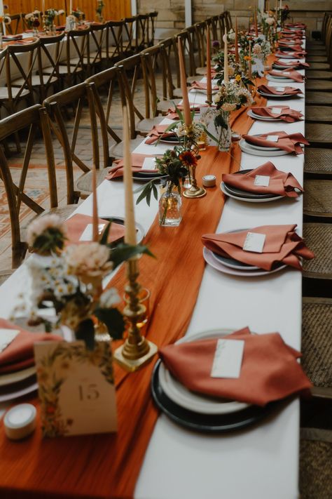 Wedding reception table decor with long burnt orange table runner and burnt orange napkins. Taper candles stand in brass candle holders & elegant Autumn flowers sit in small glass bud vases. Long trestle tables with cross back chairs. Burnt Orange Table Runner Wedding, White And Burnt Orange Wedding Table, Rustic Wedding Decor Autumn, Autumn Wedding Diy Decorations, Burnt Orange And Brown Wedding Decor, Diy Wedding Table Set Up, Burnt Orange Themed Wedding, Rustic Wedding Orange, Fall Wedding Bud Vases Centerpiece