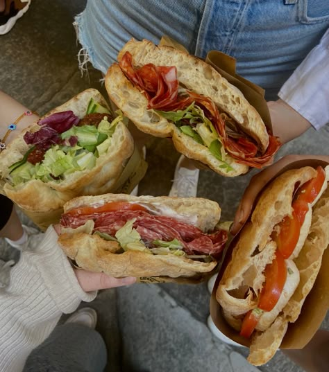 Four hands holding four different Panini with different toppings. They contain salami, tomatoes, salad, cream cheese and mozzarella. Foods In Italy, Italy Aesthetic Travel, Firenze Italy Aesthetic, Italian Food In Italy, Italy Food Aethstetic, Panini Aesthetic, Italian Food Truck, Florence Italy Aesthetic, Pasta In Italy