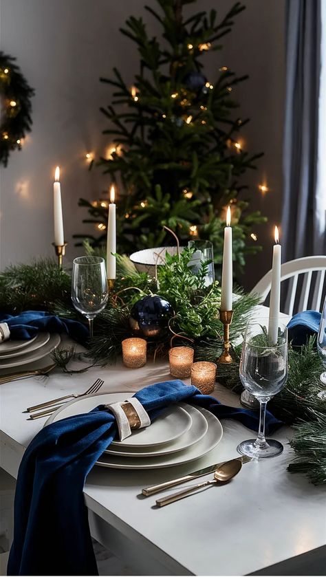 Festive dining table set with candles, greenery, blue accents, and a lit Christmas tree in the background. Midnight Blue Table Setting, Light Blue Table Setting, Christmas Table Blue, Orange And Blue Christmas, Blue Christmas Table Decor, Dark Blue Christmas Decor, Midnight Blue Christmas, Blue Christmas Table, Aesthetic Navy Blue