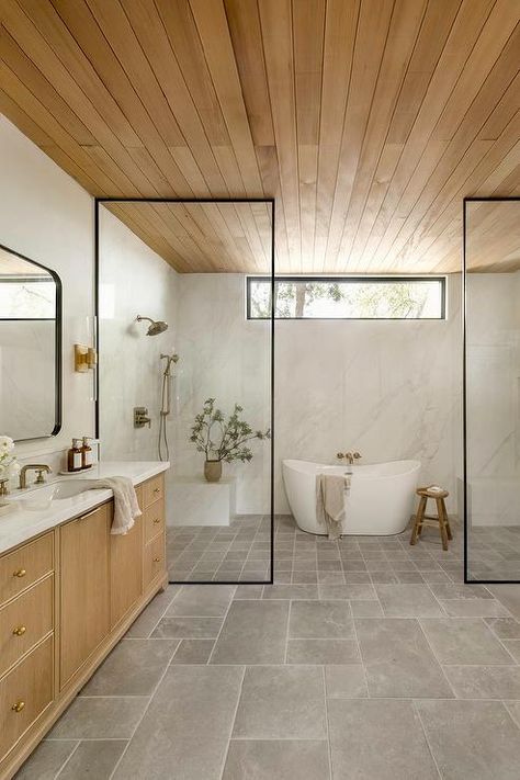 Spacious bathroom features an oval bathtub inside a walk in shower with his and hers shower benches, a clerestory window, a satin nickel shower kit on marble slab shower walls, gray grid shower floor tiles, a steel partition, a brown reeded double washstand with marble countertop with brass faucets under curved black mirrors lit by brass and glass sconces and a teak plank ceiling. Lhk Interiors, Bathroom Renovation Cost, Bad Inspiration, Trendy Bathroom, Bathroom Pictures, Grey Bathrooms, Dream Bathroom, Wet Rooms, Shower Design