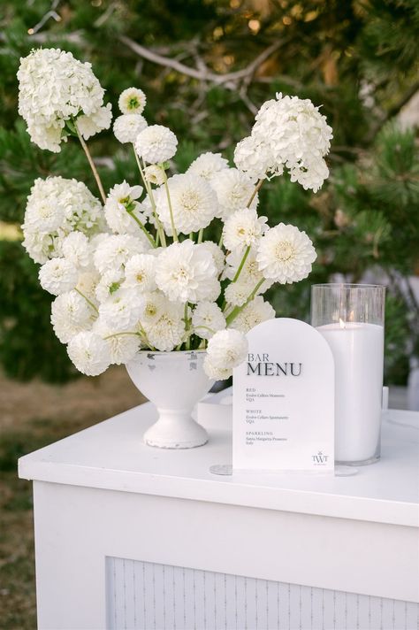 If you are planning at all white party, save this photo as decor inspiration for your event! This bar featured a white acrylic bar menu sign and elegant white floral arrangement. This is the perfect decor for a white wedding of all white party! Hampton Party Decor, All White Arrangements, Welcome Ceremony Decorations, White Alter Flowers, Simple Wedding Cocktail Table Decor, Chic Table Numbers Wedding, Diy Flower Pedestal Wedding, White Garden Party Decoration, White Floral Wedding Reception