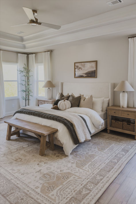 Neutral bedroom with warm wood tones, an upholstered bed, soft bedding, and a patterned area rug. The space includes natural accents like a wooden bench, woven baskets, and greenery. A pair of pottery lamps sit on wood nightstands, framed by white curtains that add to the light, airy feel. The green throw blanket and subtle green accents bring warmth and depth, while the gold ceiling fan and framed art complete the Pottery Barn-inspired look. Pottery Barn Bedroom Master, Bedroom Pottery Barn, Light Airy Bedroom, Gold Bed Frame, Pottery Barn Bedrooms, Cozy Neutral Bedroom, Pottery Lamps, Gold Ceiling Fan, Wood Nightstands