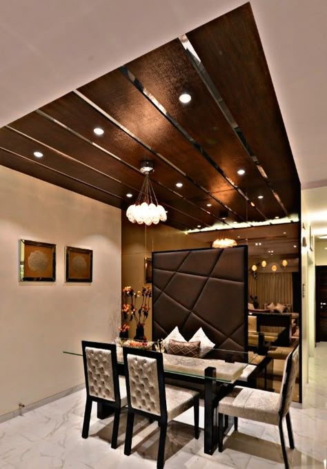 This dining room is an extension to living room wherein the lighting plays the pivotal role to divide two areas, which is enhanced even more by the brown tinted mirror reflecting it along with the leather finish backdrop having abstract line play, adding to the richness. Mirror Ceiling Living Room, Wooden Ceiling Design Dining Area, Brown Mirror Wall Interior Design, Brown Ceiling Living Room, Veneer Ceiling Design Living Room, Brown Tinted Mirror, Mirror Ceiling Design, Wooden Ceiling Design Living Room, Wooden False Ceiling Design