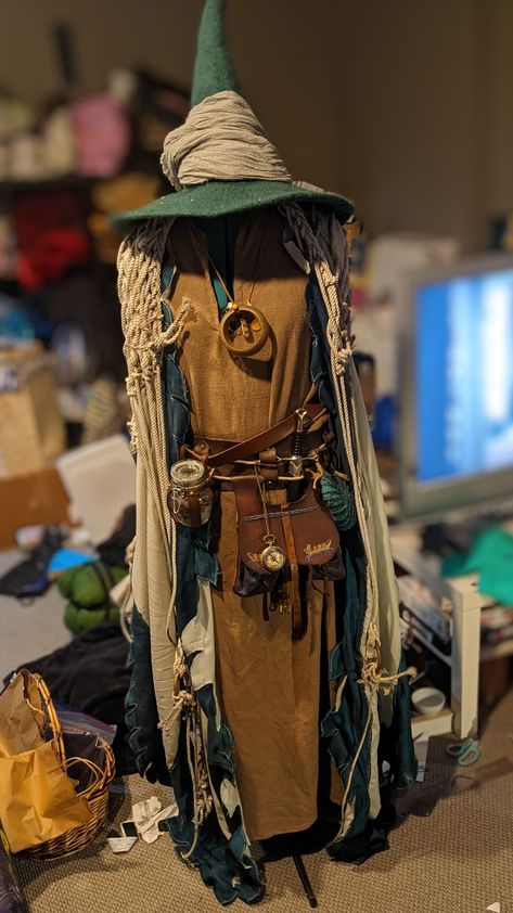 A beach wizard inspired costume on a mannequin, the base garment is a sandy coloured linen tunic with multiple belts around the waist. There is a jar of beach glass, a compass, a medieval bollock bag and dagger, and a crocheted seashell all hanging off the waist. A floor length mint robe with shimmery dark teal borders with dagging and floor length angel sleeves is worn under what appears to be an old fishing net worn as a shawl. Stop is a green wool wizard hat wrapped with gray trim Crochet Wizard Robe, Ren Faire Wizard Costume, Forest Wizard Costume, Wizard Cosplay Men, Wizard Ren Faire, Wizard Costume Women, Wizard Costume Ideas, Ren Faire Wizard, Wizard Aesthetic Fashion