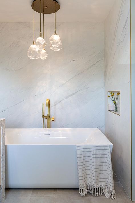 This modern bathroom has a freestanding white bathtub that's recessed into its own alcove. A sculptural multi-light pendant lamps with glass geometric shades, hangs above the bath, highlighting the design. #Bathtub #ModernBathroom Bathtub Lighting, White Bathtub, Bathroom Lighting Design, Bathroom Lighting Ideas, Brass Shower Head, Bathroom Lights, Bathroom Design Inspiration, White Chandelier, Wood Framed Mirror