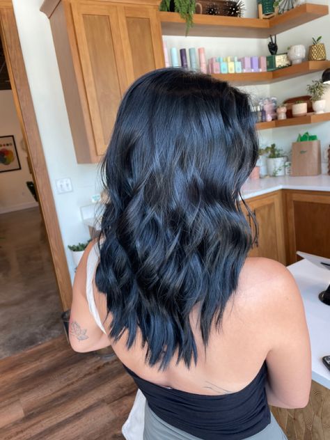 Girl with curled, blue-black hair standing with her back turned to the camera. She’s standing in the foreground in a hair salon with wooden cabinets behind her. She is wearing green pants and a black halter top with a cream corduroy bag on her left shoulder. Black Hair Color With Blue Tint, Black Hair With Tint Of Blue, Black Saphir Hair, Blue Tone Black Hair, Ion Blue Black Hair Color, Blue Black Hair Layers, Jet Black With Blue Tint, Subtle Blue Black Hair, Cool Tone Black Hair Color