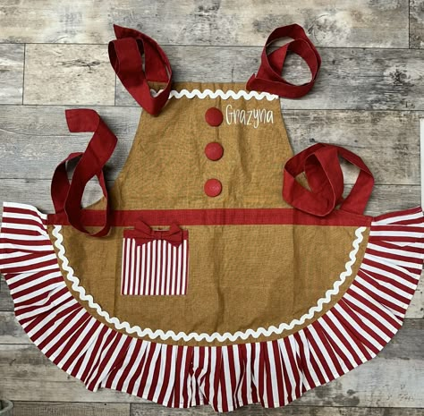 ADULT GINGERBREAD APRON Personalized with Chef and the your name on left chest Search for the matching Childs Gingerbread Apron  Details Adult light brown gingerbread apron with three large buttons. Pocket and ruffle are red and white stripe and pocket has a red bow. Made of fabric and buttons. 27"H X 37"W When Ordering Please Be Sure To Leave The Following In Notes Section: Name in which you would like placed on apron Font Style And thread color Please don't hesitate to request a preview of the font before ordering to make sure you like it. Holy Stitchin will not replace items that are personalized if you don't like a font you request. Gingerbread Apron Diy, Cute Apron Pattern Free, Diy Christmas Apron, Christmas Aprons Diy, Christmas Aprons Ideas, Princess Apron Pattern, Aprons Diy, Apron Diy, Gingerbread Apron