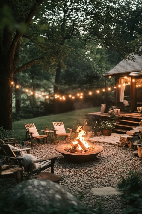 Cozy outdoor fire pit area with string lights, surrounded by wooden chairs and lush greenery. Fire Pit With Rocks Natural Stones, Fire Outside Ideas, Rustic Farmhouse Backyard, Modern Backyard Fire Pit Ideas, Patio Area With Fire Pit, Outdoor Fire Pit Lights, Home With Backyard, Garden Design With Fire Pit, Backyard Gathering Space