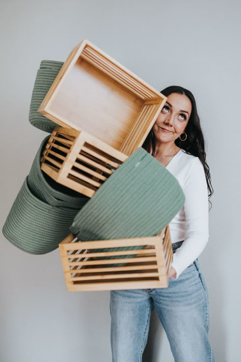 A woman with dark hair wearing a white long-sleeve shirt and blue jeans is holding a stack of balancing green fabric baskets and wooden crates while look up and to the right. Staging Branding Photos, Organizer Branding Shoot, Professional Home Organizer, Professional Organizer Photoshoot, Personal Organizer Ideas, Linen Photoshoot, Professional Organizer Business, Branding Headshots, Co Branding