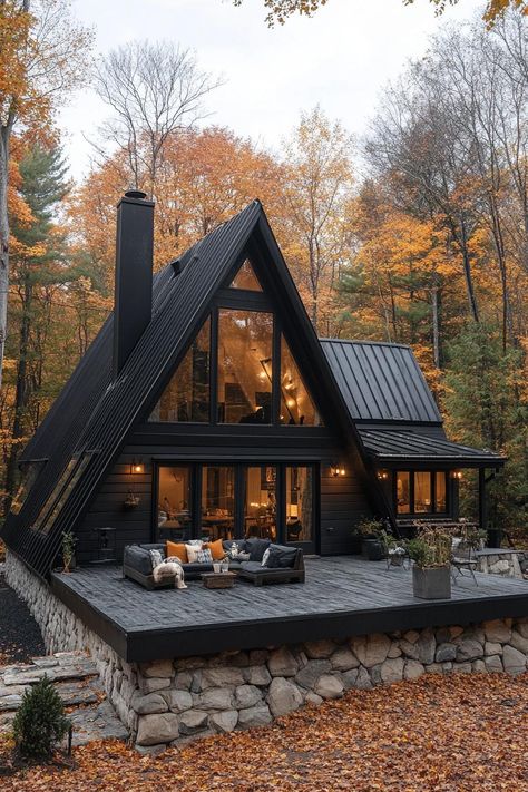 Large a frame house with charred wood siding black metal roof with black chimney stone foundation with front deck with outdoor furniture house. Check out all of these stunning and chic dark A-frame houses that pack a ton of charm into a small forest retreat. Dark Exterior Cabin, Forest Small House, A House In The Forest, Dream Forest House, Dark Stain Log Cabin Exterior, A Frame Tiny House Floor Plans, Metal A Frame House, Black Gothic House Exterior, Gothic A Frame House