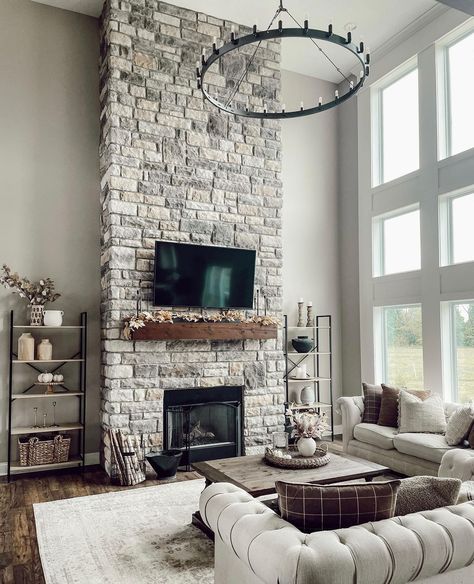 A flat panel TV is mounted to a gray stone two story fireplace above a stained wood mantel. The TV faces a light gray Chesterfield sofa and wood coffee table lit by a black wagon wheel chandelier. Stone Tile Fireplace, Shiplap Living Room, Stained Wood Beams, Farmhouse Fireplace Ideas, Grey Stone Fireplace, Stained Wood Trim, Two Story Fireplace, Fireplace With Tv, Stone Fireplace Wall