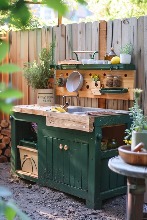 Outdoor wooden kitchen with green cabinets, potted plants, and cooking utensils against a wooden fence background. Palette Mud Kitchen, Mid Kitchen, Diy Pallet Mud Kitchen Outdoor Play, Mud Kitchen On Fence, Forest School Mud Kitchen, Outdoor Mud Kitchen, Wood Pallet Mud Kitchen, Kitchen For Kids, Outdoor Play Kitchen