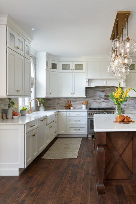 White kitchen with stone backsplash, glass door cabinets, under cabinet outlets, gold hardware, island chandelier, and stained island on wood floor. White Kitchen With Walnut Island, Brown And White Cabinets Kitchen, White Cabinets Gold Hardware Kitchen, Kitchen With Stone Backsplash, Gold Hardware Kitchen, Tan Kitchen Cabinets, Under Cabinet Outlets, White Kitchen Dark Floors, Stained Island