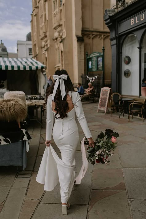 Bride in open back detailed long sleeve wedding dress with buttons down the middle and large white bow hair accessory walking to her wedding first look White Bow In Hair Wedding, Bridal Hairstyle With Bow, Hair Bow Bride, Hair With Bow Wedding, Half Up Hair With Bow Wedding, White Bow Aesthetic, White Bow Wedding Hair, Bridal Bow Hairstyle, Bridal Shower Hair With Bow