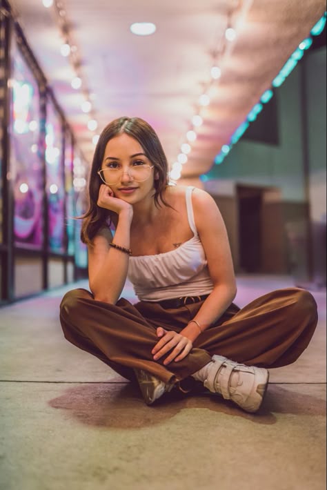 A night Photography photo of a beautiful girl with brown hair wearing a white tank top, brown pants, and white shoes, with glasses sitting down criss cross in front of bright lights Girl Sit Reference, Sitting Poses Criss Cross, Criss Cross Sitting Pose, Sitting Pose Reference Criss Cross, Crisscross Sitting Pose, Women Sitting Cross Legged, Sitting Cross Legged On Floor, Pose Reference Criss Cross, Cross Legged Sitting Pose