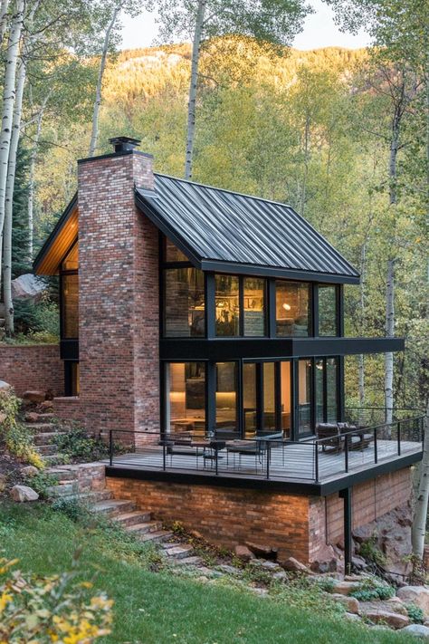 Small modern mountain house facade with varying shade brick wood and black steel accents slate tile roof with a brick chminey raised stone deck in mountain forest clearing. Check out all of these charming small mountain houses that blend style with nature for an affordable dream retreat. Mountain Top House Plans, Glass Box House Architecture, Narrow Hillside House Plans, Tiny House On A Hill, House On A Steep Slope, Small House On Hillside, Small Homes With Lots Of Windows, Cabins By The River, House On A Creek