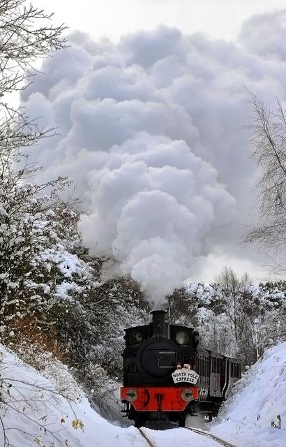 a train traveling down tracks next to snow covered ground and trees with steam pouring out of the top