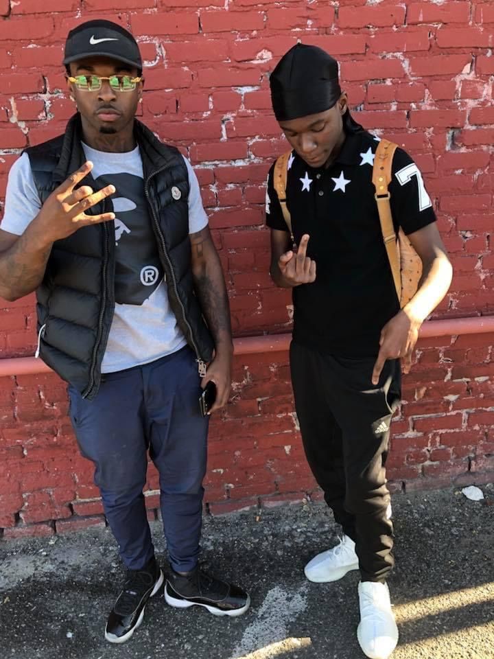 two young men standing in front of a red brick wall with one pointing at the camera