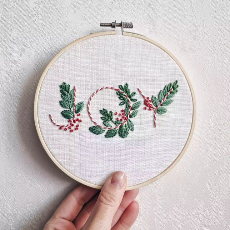 a hand is holding up a cross stitch hoop with holly leaves and berries on it