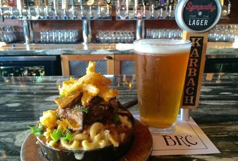 a wooden plate topped with pasta and meat next to a tall glass filled with beer