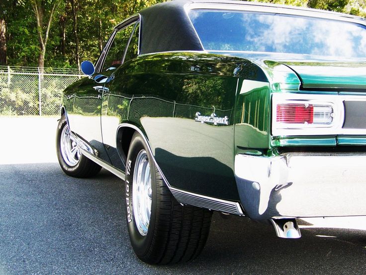 the rear end of a green car parked in a parking lot with trees behind it