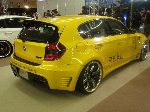 two yellow cars parked next to each other in a showroom with people looking at them