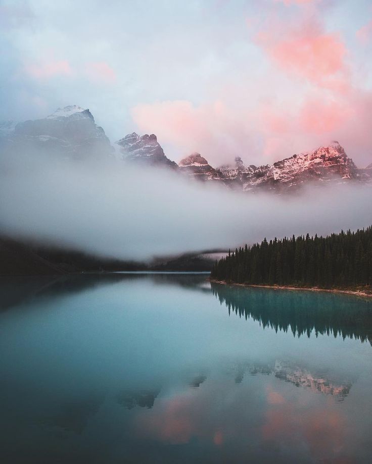 a lake surrounded by mountains with clouds in the sky and trees around it at sunset