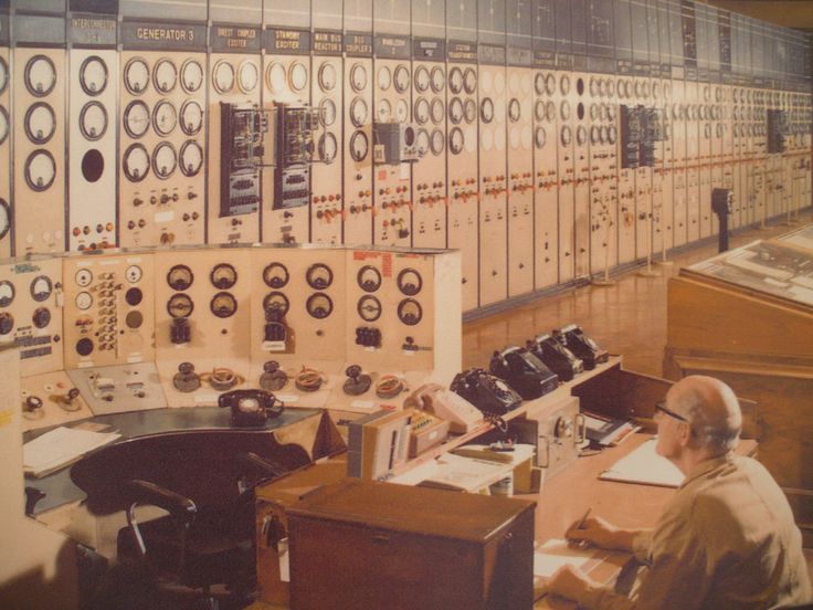 an older man sitting at a desk in front of many control boards