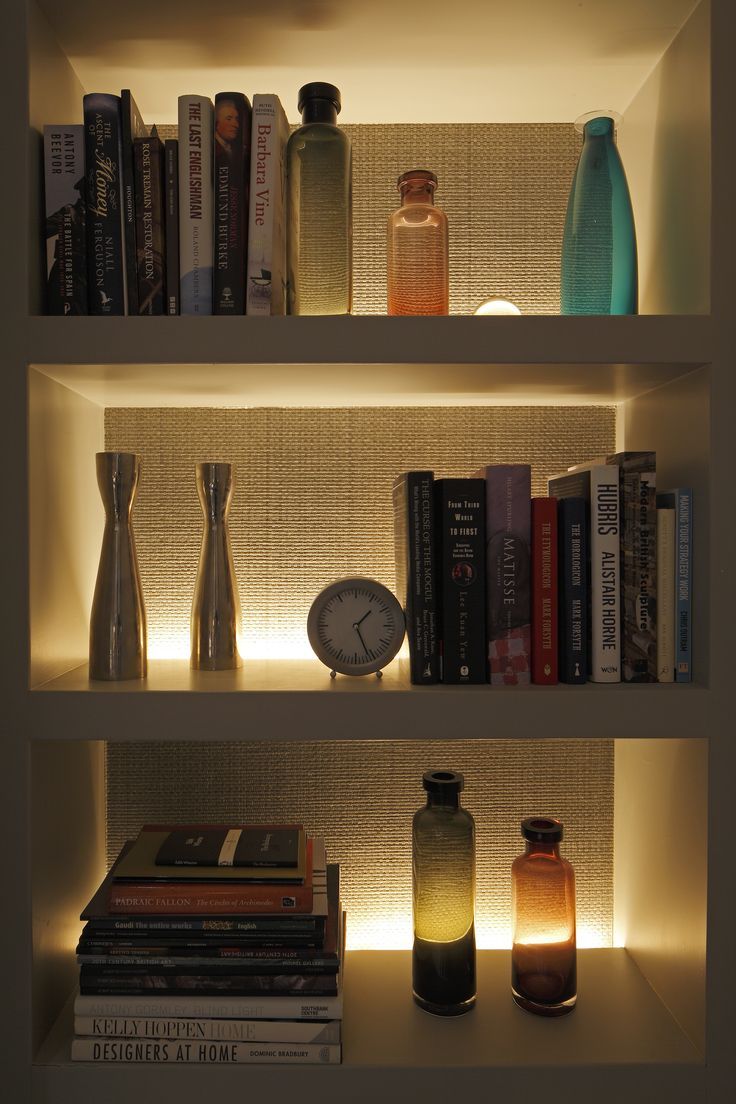 some books and bottles on a shelf with lights in the corner behind them, along with a clock