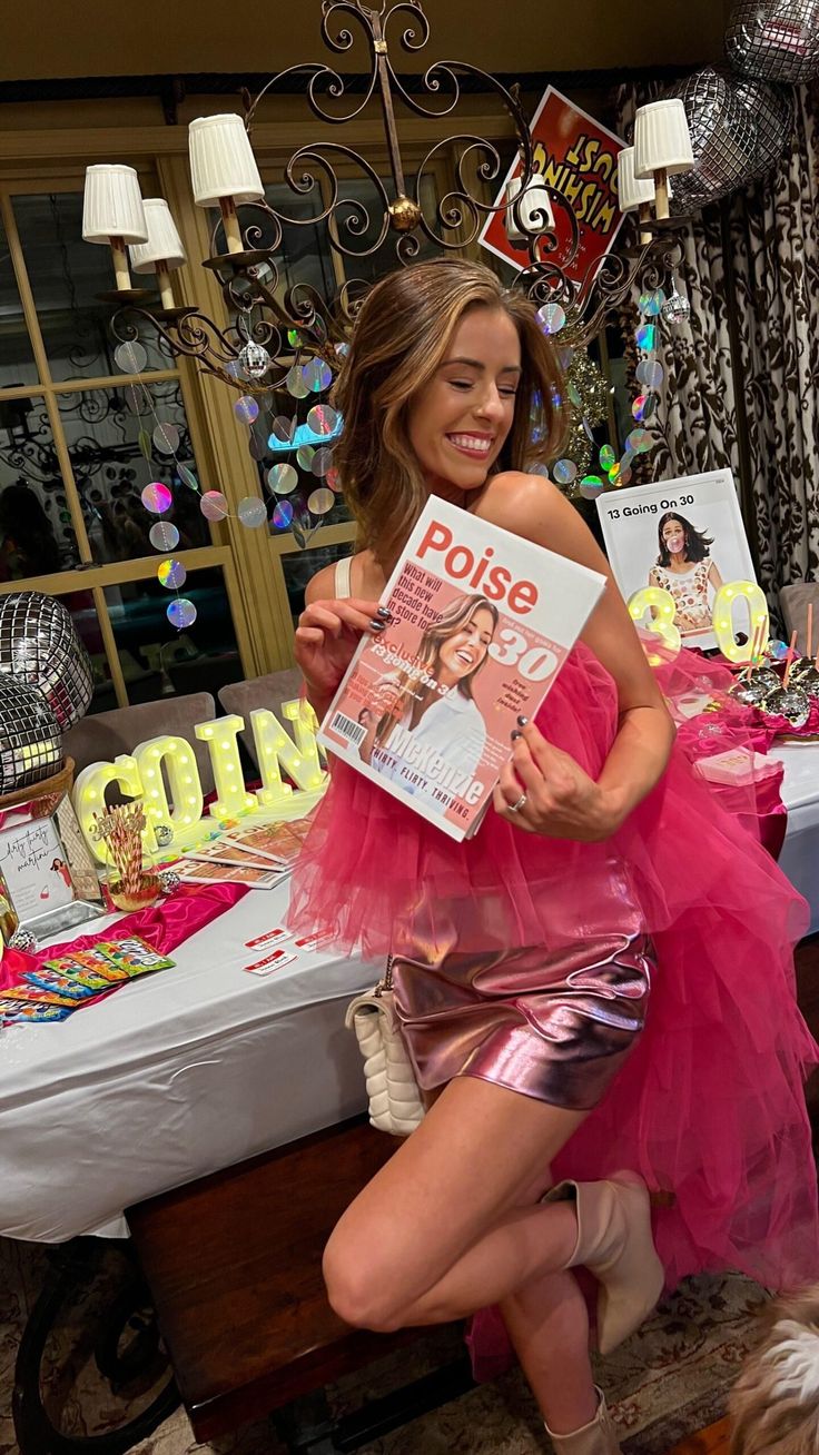 a woman in a pink dress sitting on a table holding a magazine and posing for the camera