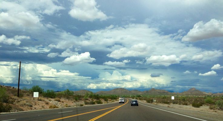 cars driving down an empty highway with mountains in the backgrouund and clouds in the sky