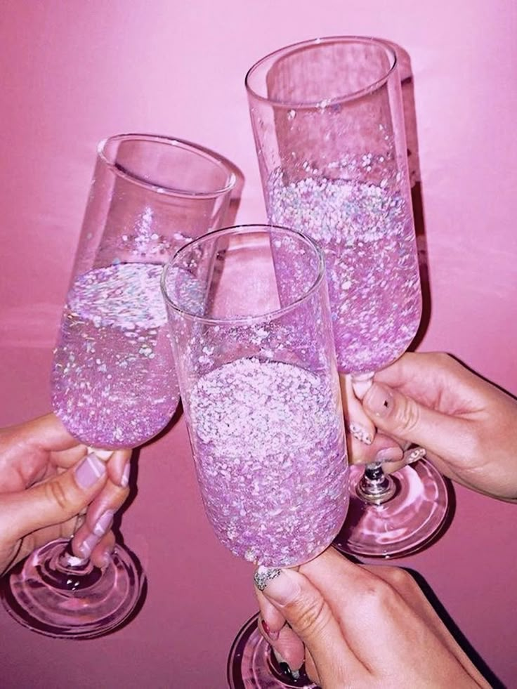 three people holding up wine glasses filled with pink liquid and silver flakes on the rims