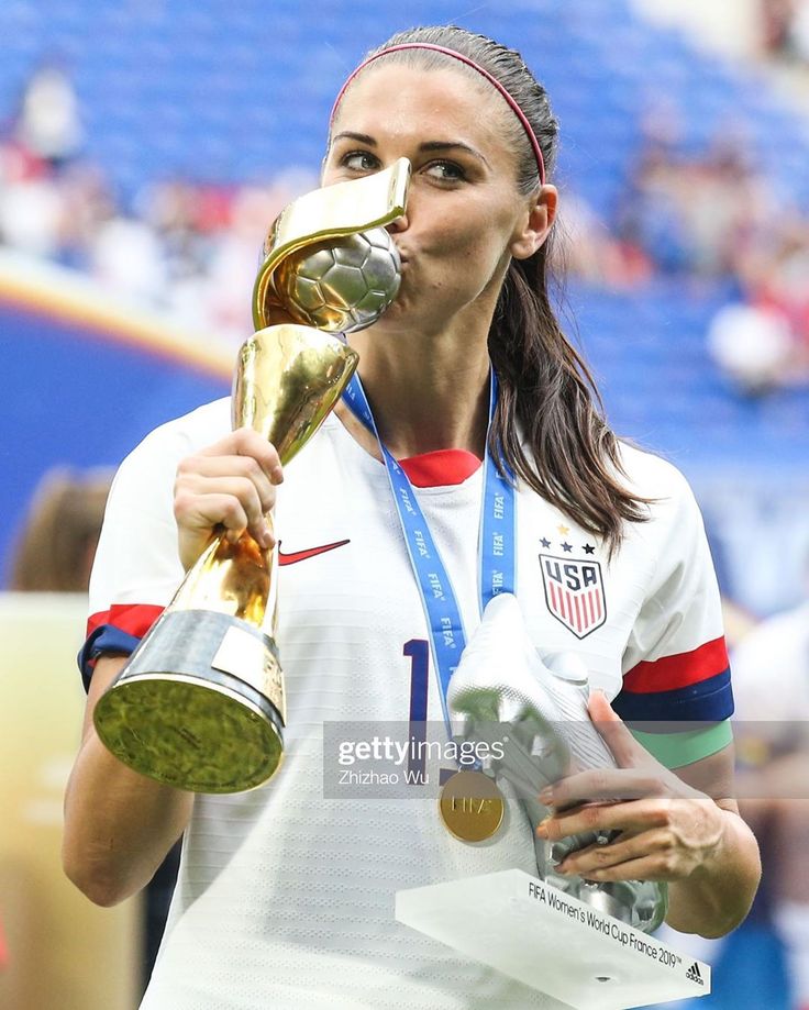 a female soccer player holding a trophy and blowing it into her mouth while standing on the field