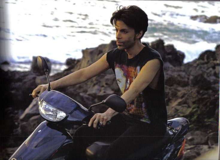 a man riding on the back of a blue motorcycle next to the ocean and rocks