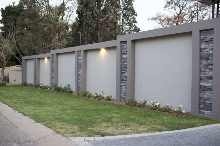 a fenced in area with grass and bushes next to the wall that has lights on it