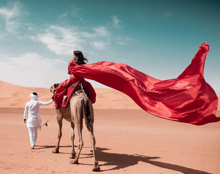 a man in white is walking with a camel and a red cloth over his head