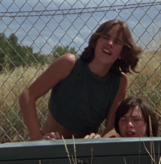 two people standing behind a fence in front of tall grass and bushes, with one person sticking his head over the fence