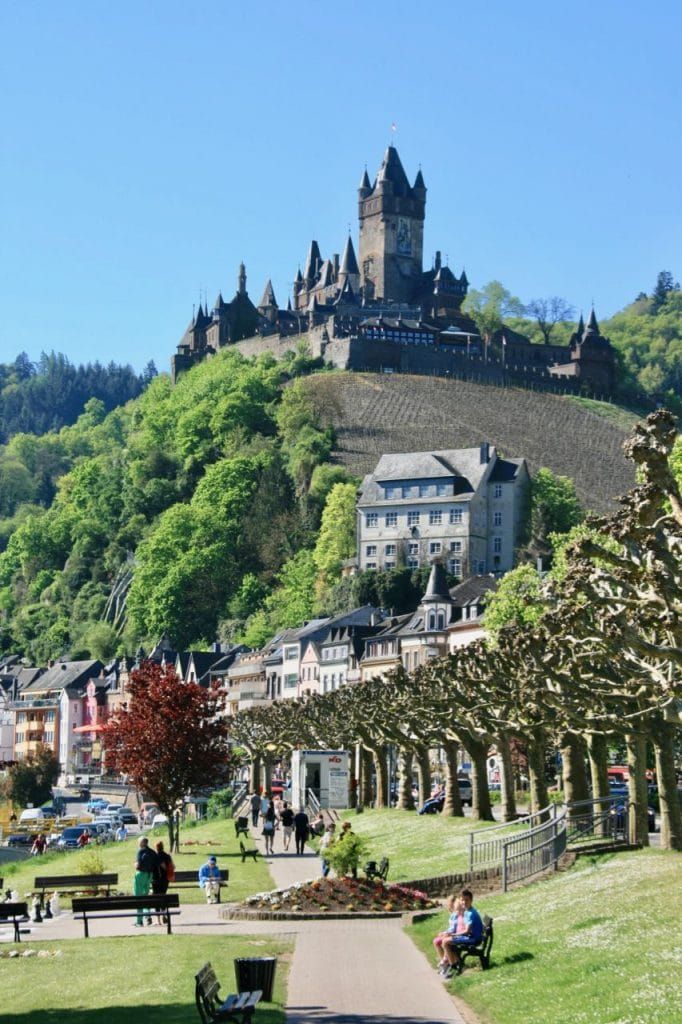 people are sitting on benches in the grass near a large castle like building that is perched on top of a hill