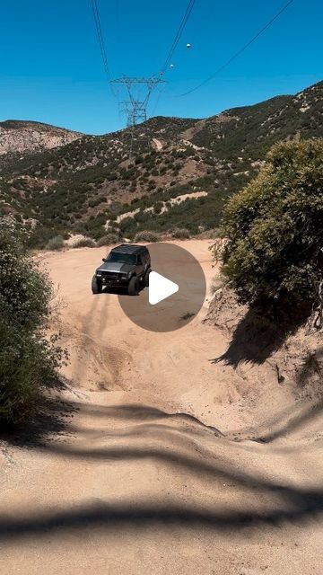 a car driving down a dirt road in the middle of some bushes and trees with power lines above it