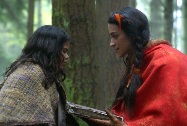 two women dressed in native american clothing standing next to each other near a tree trunk