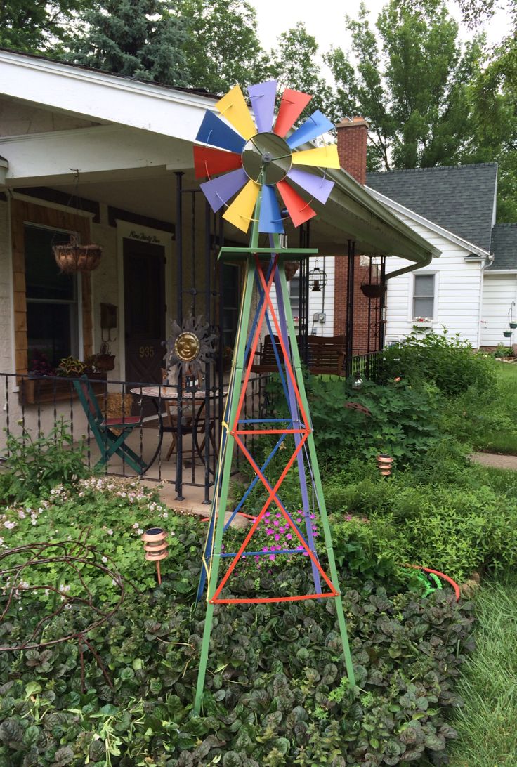 a colorful windmill sitting in the middle of a garden next to a house and bushes