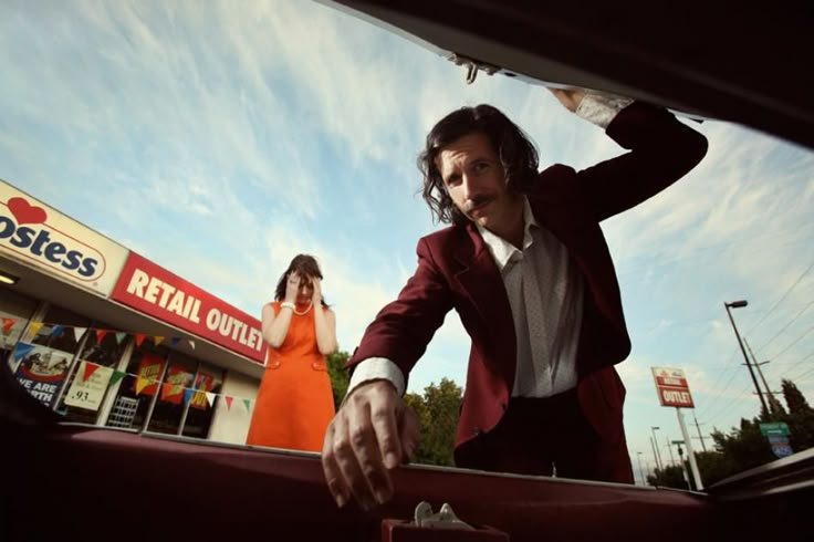 a man in a red suit and white shirt standing next to a woman with her hand on the hood of a car