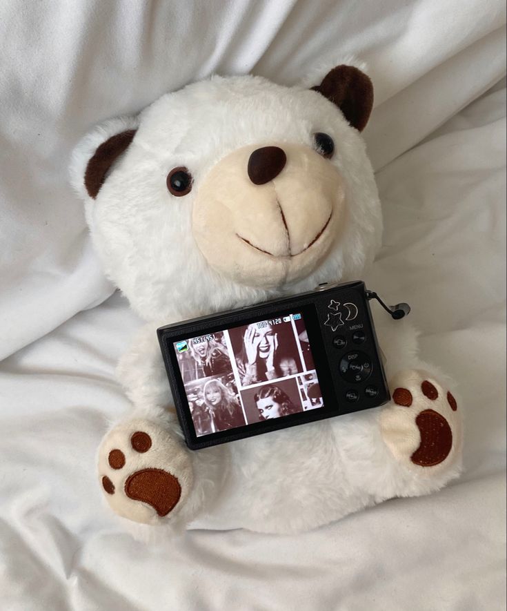 a white teddy bear sitting on top of a bed holding a cell phone in its paws