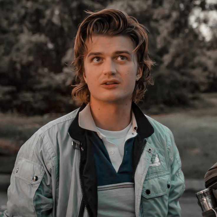 a young man with long hair standing in front of trees and looking at the camera