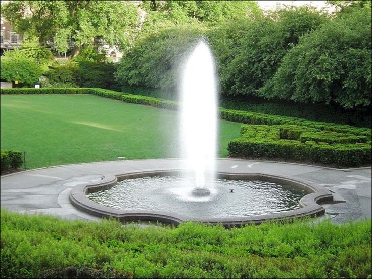 a water fountain is in the middle of a circular area with hedges and trees around it