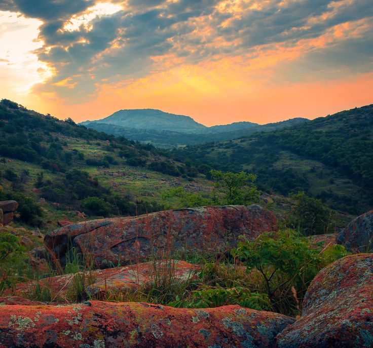 the sun is setting over some rocks and trees with mountains in the backgroud