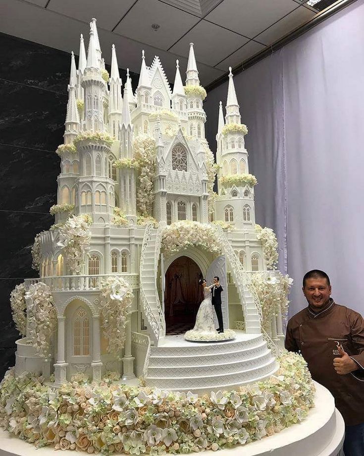 a man standing next to a large cake with white frosting on it and a castle in the middle